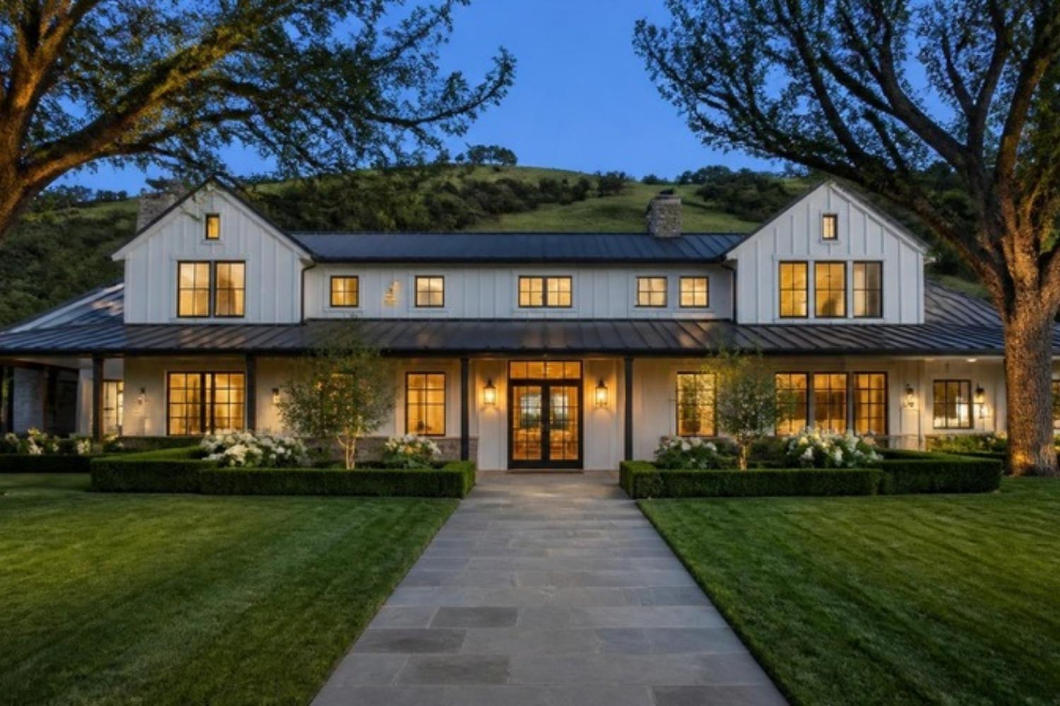 A wide stone front walk crossing a green lawn to the lit entry of a modern farmhouse estate at dusk